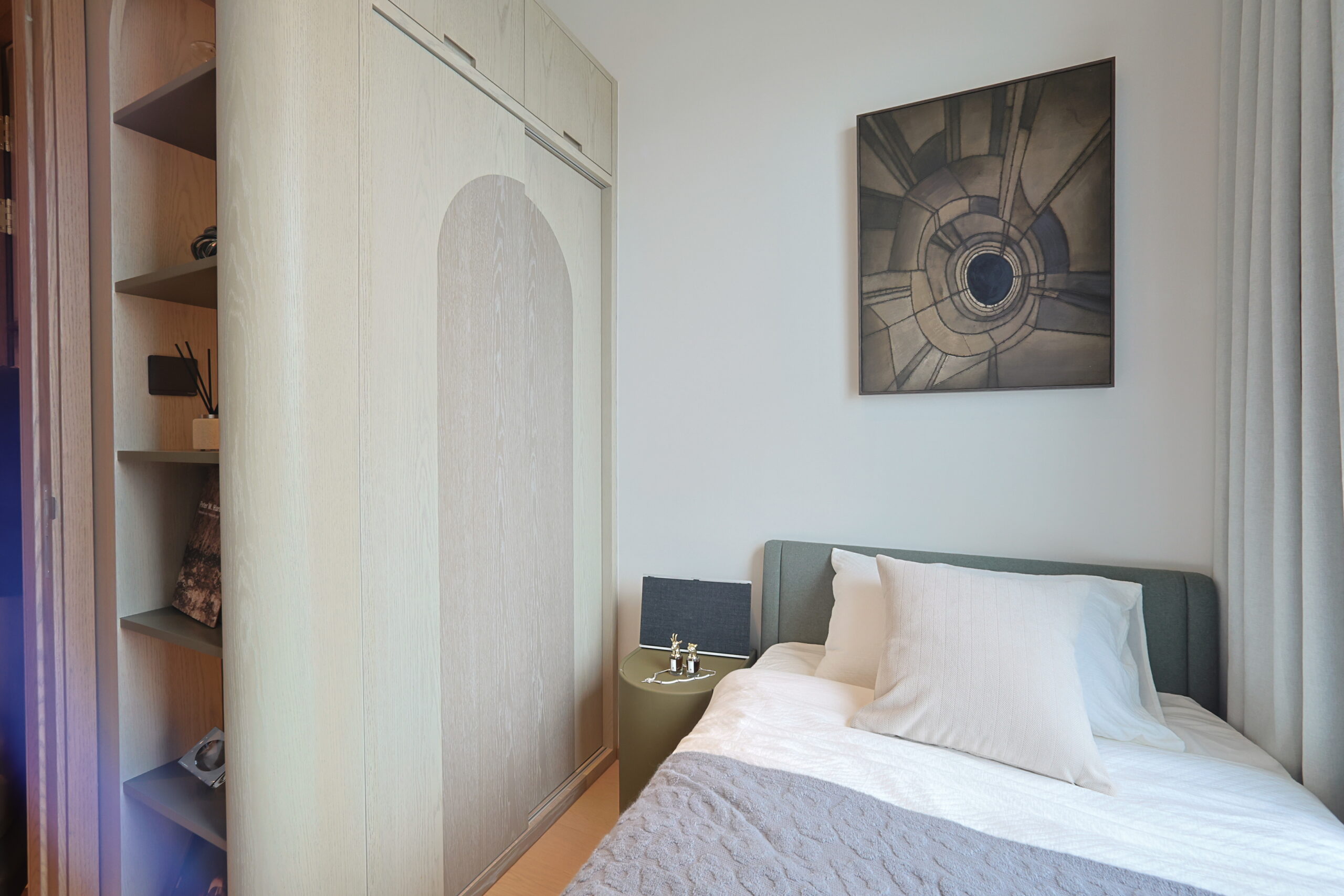 Hotel-style bedroom with a tall light-wood wardrobe on the left, bed on the right, and a circular abstract art piece on the wall.