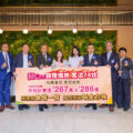 Group of professionals in business attire pose with a large pink banner in a modern lobby backdrop with green wall art behind them signaling a promotional event