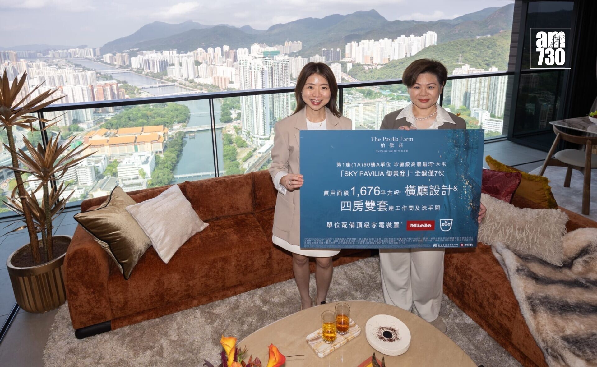 Two women in beige outfits hold a large blue promotional board inside a high-rise living room, with a panoramic city skyline and river behind them; AM730 logo appears in the top-right corner.
