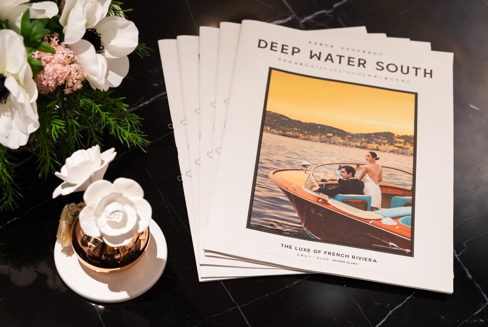 Stack of magazines titled Deep Water South; cover shows a couple in a wooden boat on sunset water, with flowers on a marble surface nearby.