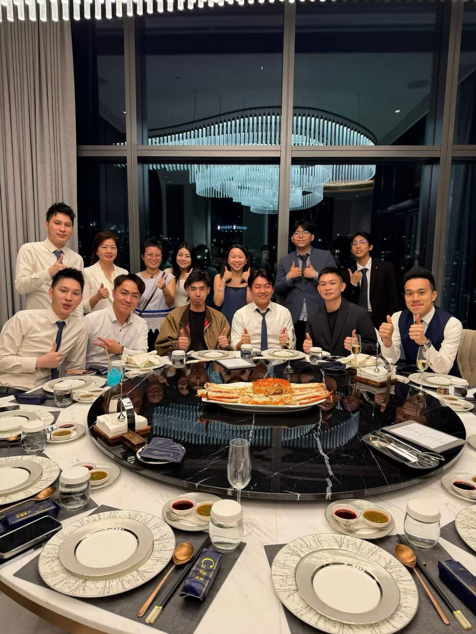 Group of colleagues in formal wear posing with thumbs up around a round dining table, centered by a seafood platter.