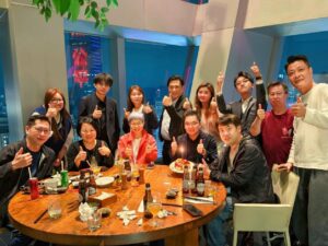 A group of friends and colleagues sit around a round table in a restaurant giving thumbs up as a birthday cake with a lit candle sits in the center