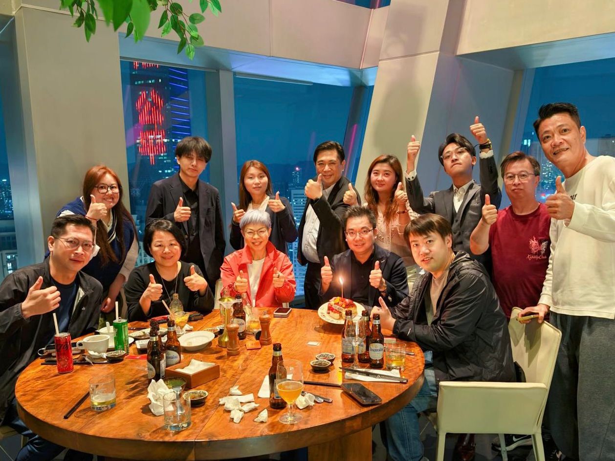 A group of friends and colleagues sit around a round table in a restaurant, giving thumbs up as a birthday cake with a lit candle sits in the center.