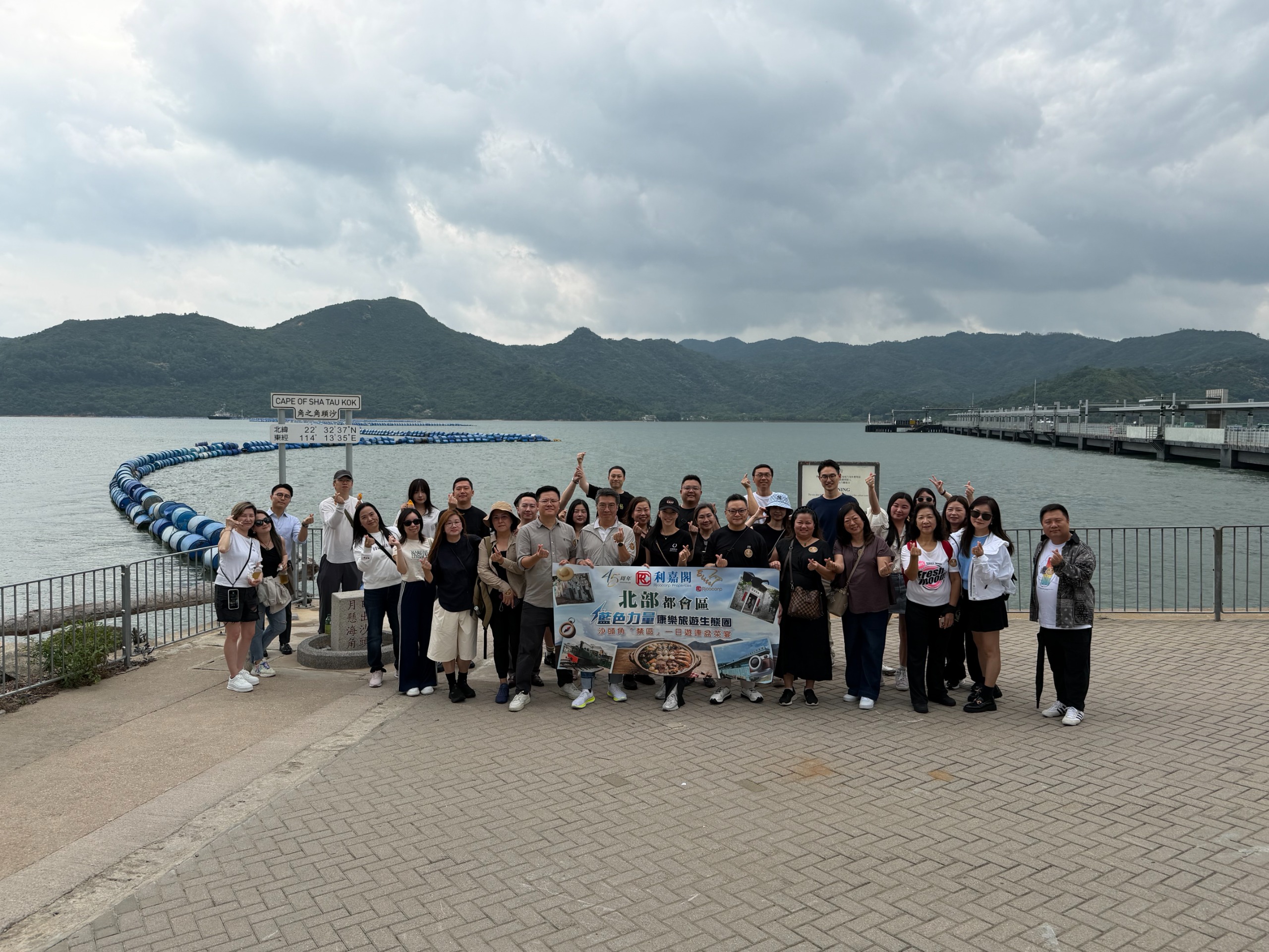 Large group of people posing together on a waterfront promenade, holding a banner, with hills and cloudy sky behind them.