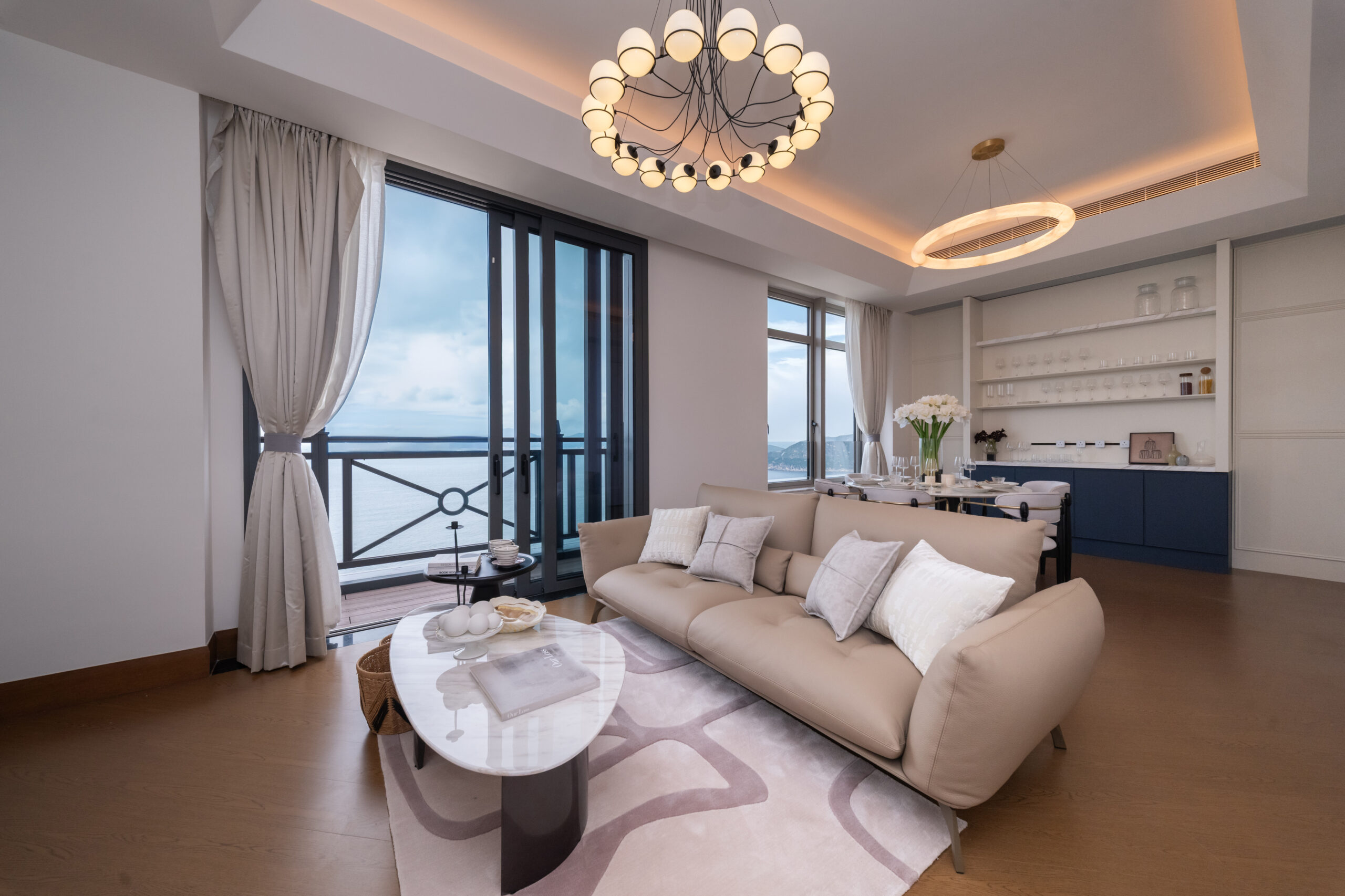 Bright, open living room with beige sectional sofa, white pillows, and a glass-top coffee table facing large windows and balcony doors. Italianate chandelier overhead.