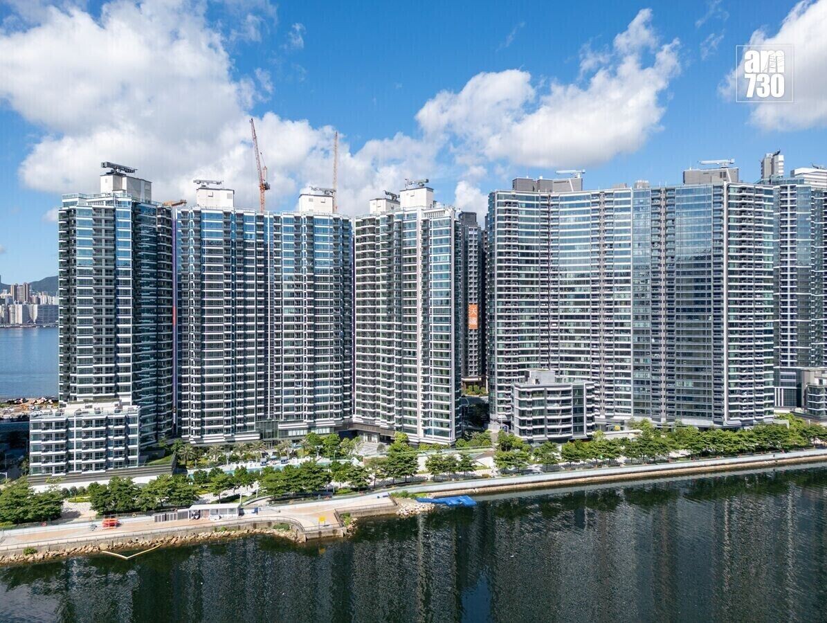 Row of modern glass apartment towers along a river, with trees and a promenade in front and blue sky.