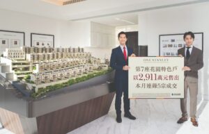 Two men in business suits pose in a real estate showroom holding a large sign beside a detailed architectural model of a residential development