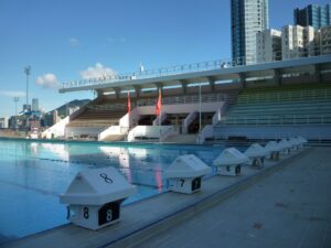 Outdoor swimming pool with white starting blocks numbered 68 along the edge and empty spectator stands urban skyline in the background
