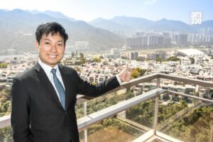 Smiling man in a suit on a balcony giving a thumbs up with a cityscape and distant mountains behind him