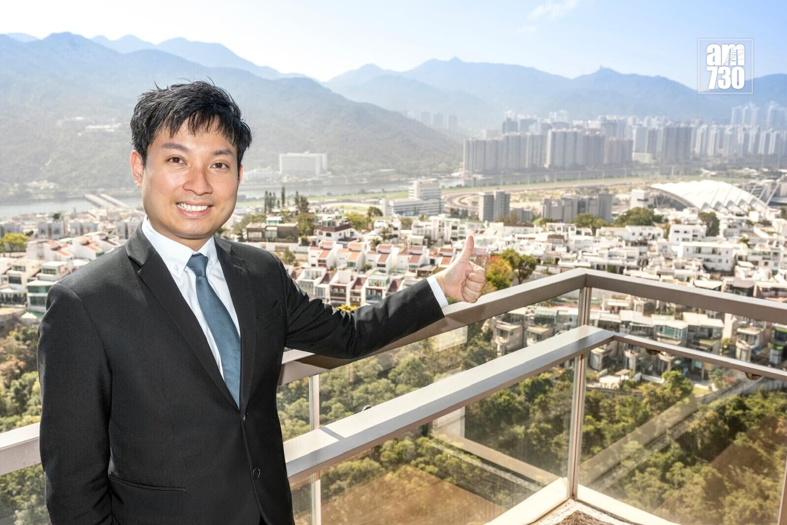Smiling man in a suit on a balcony giving a thumbs up, with a cityscape and distant mountains behind him.
