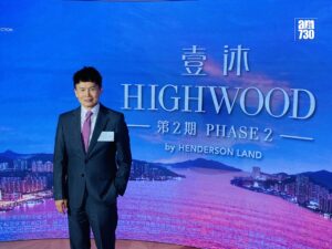 Man in a dark suit and pink tie standing in front of a blue backdrop with HIGHWOOD branding and Chinese characters at a conference