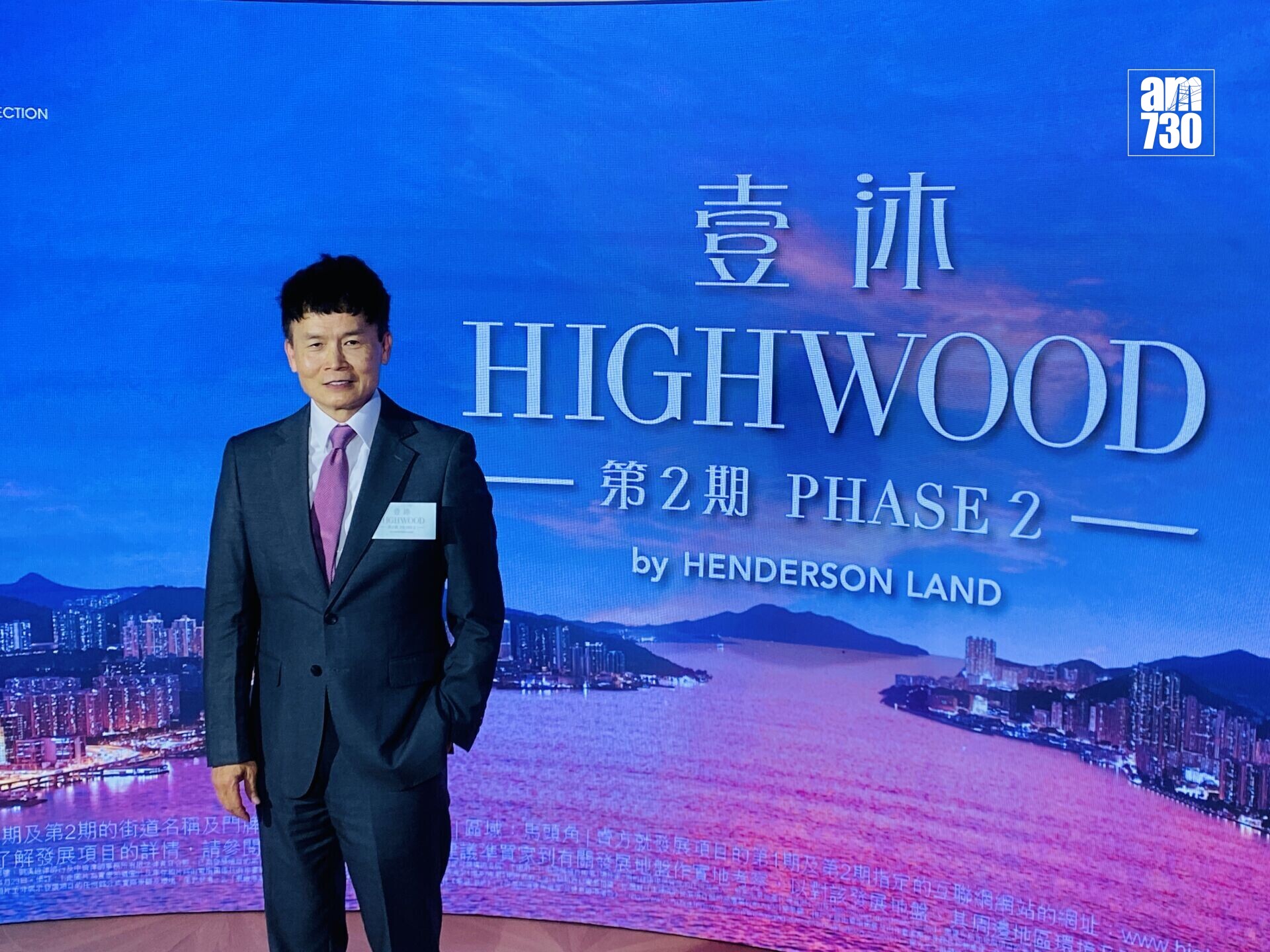 Man in a dark suit and pink tie standing in front of a blue backdrop with HIGHWOOD branding and Chinese characters, at a conference.