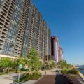 Row of glass fronted apartment towers along a waterfront promenade with trees and a blue sky