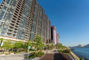 Row of glass fronted apartment towers along a waterfront promenade with trees and a blue sky