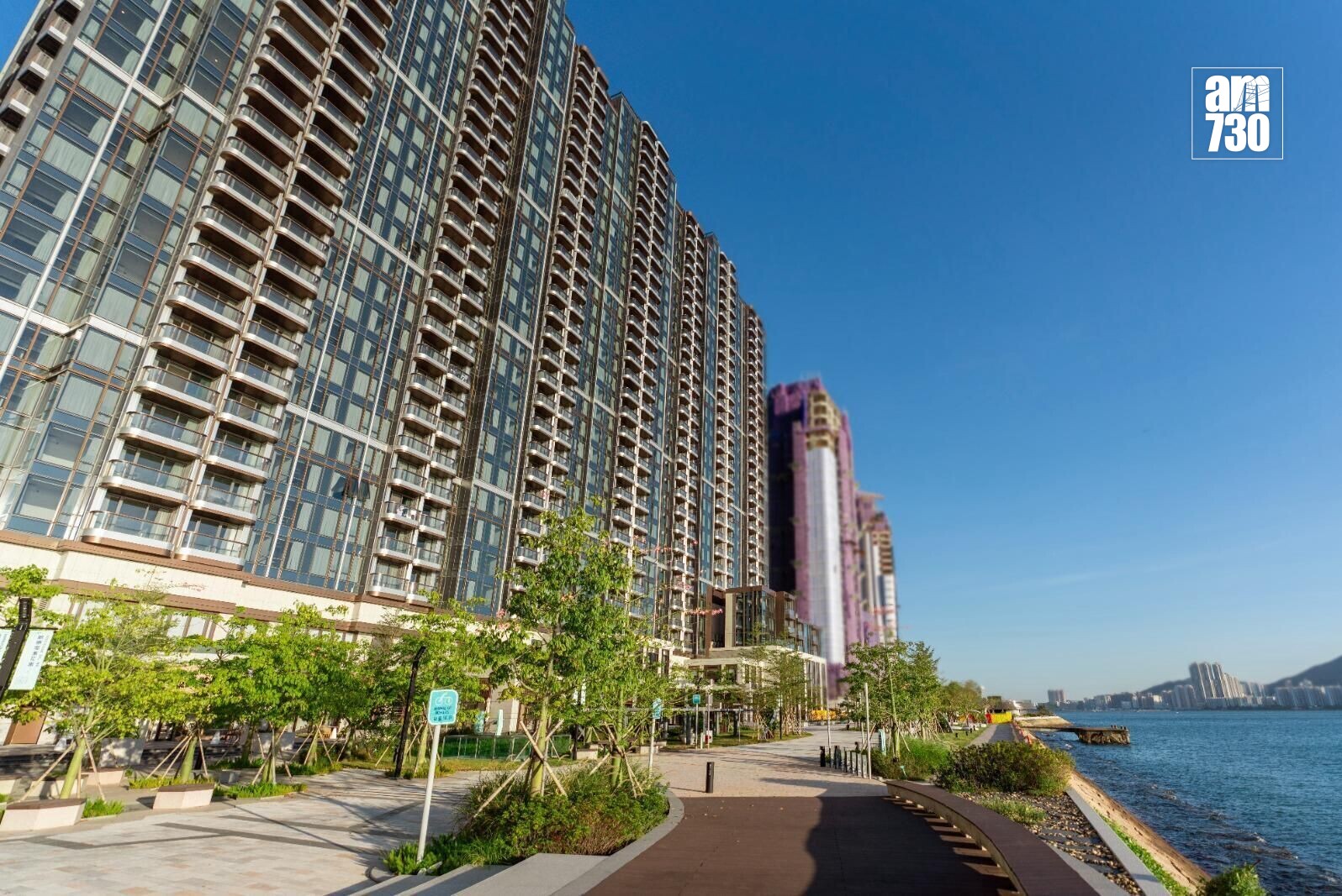 Row of glass residential towers along a waterfront promenade under a clear blue sky, AM730 logo in the top-right.