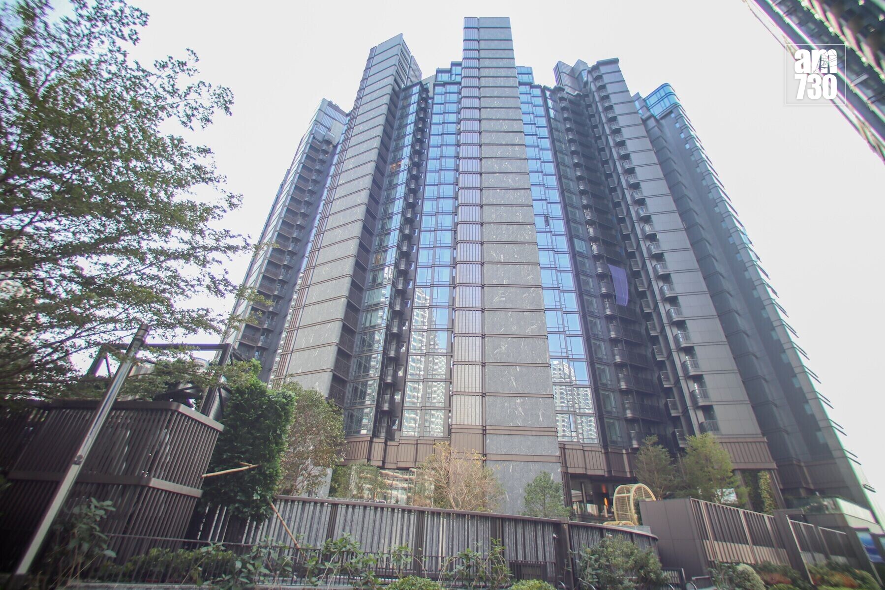 Tall modern glass-and-concrete skyscraper viewed from the street, with trees in the foreground and a pale sky behind.