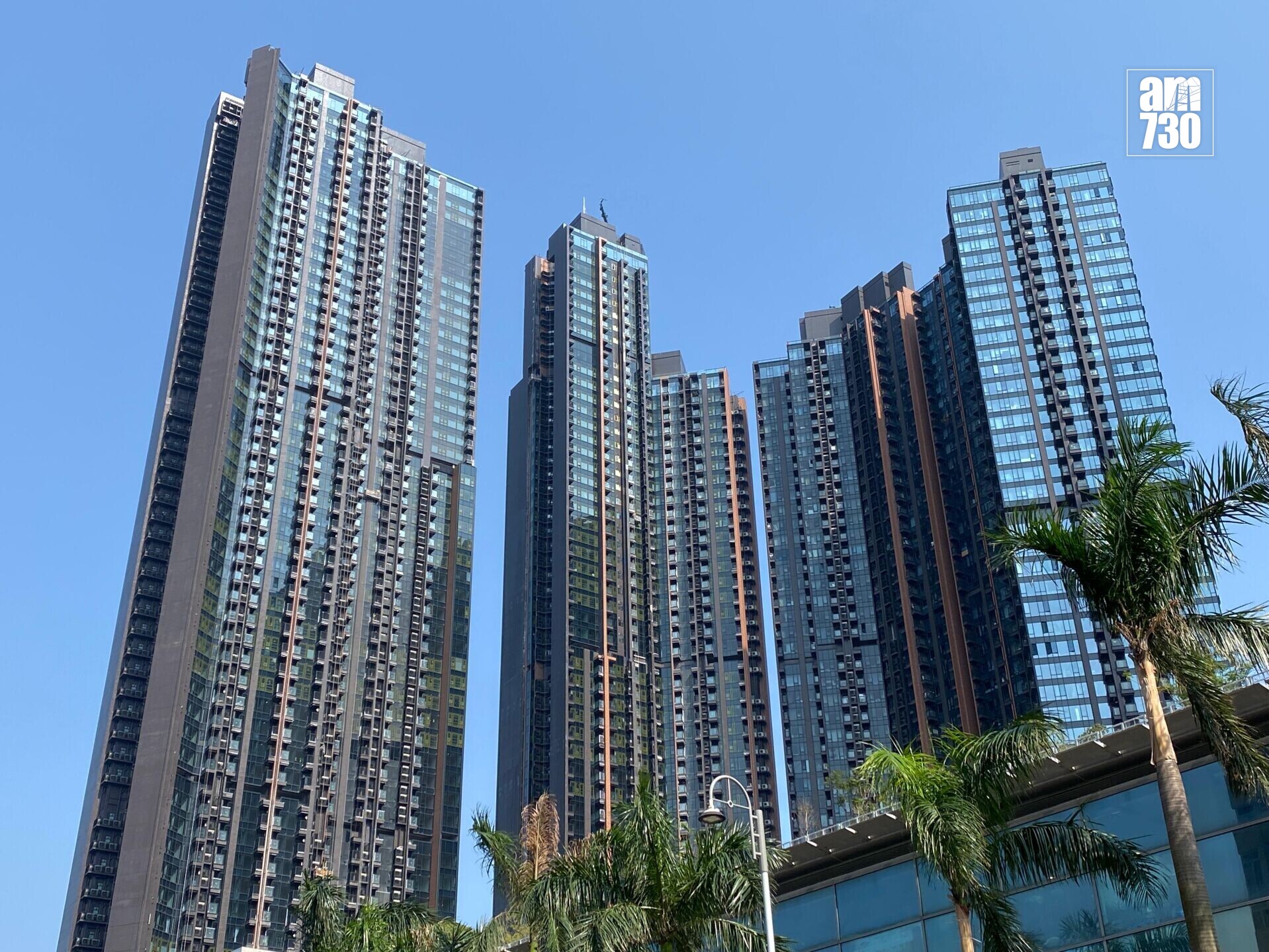 Cluster of tall glass-clad office towers reaches into a clear blue sky, with palm trees in the foreground and an AM730 logo in the top-right corner.