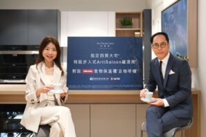 Two professionals a woman in cream and a man in a blue suit sit with coffee cups beside a large blue sign with Chinese text and logos in a modern kitchen showroom