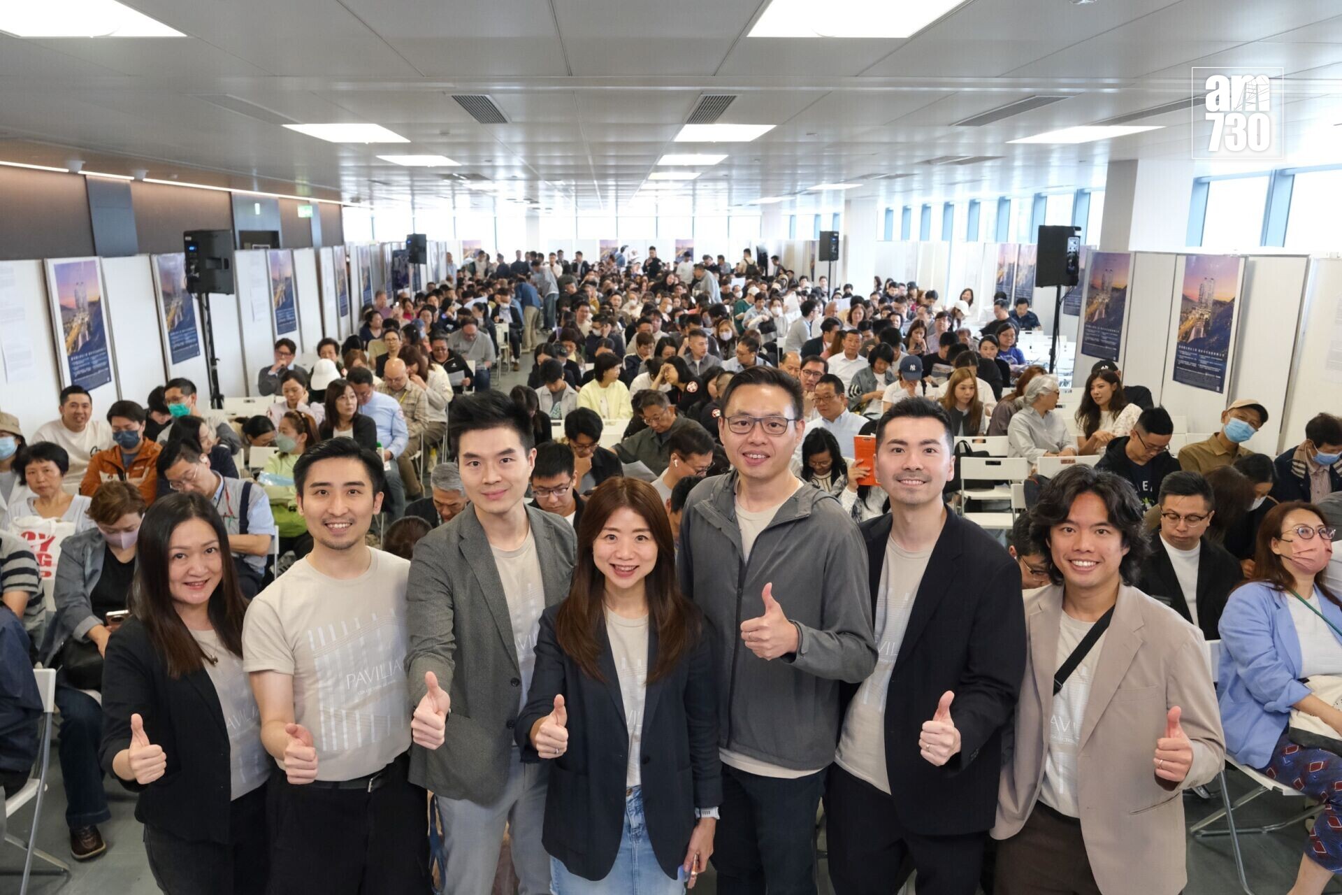 Group of people at a conference posing for a photo, front row giving thumbs up with a crowded audience behind them in a well-lit hall with poster boards along the walls.
