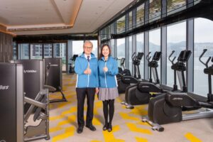 Two staff members in blue jackets posing and giving thumbs up in a bright gym with treadmills by large windows