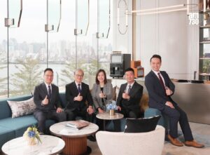 Five professionals in suits sit on a blue sofa in a bright lobby giving thumbs up with a city skyline seen through large glass panels and an AM730 logo in the corner