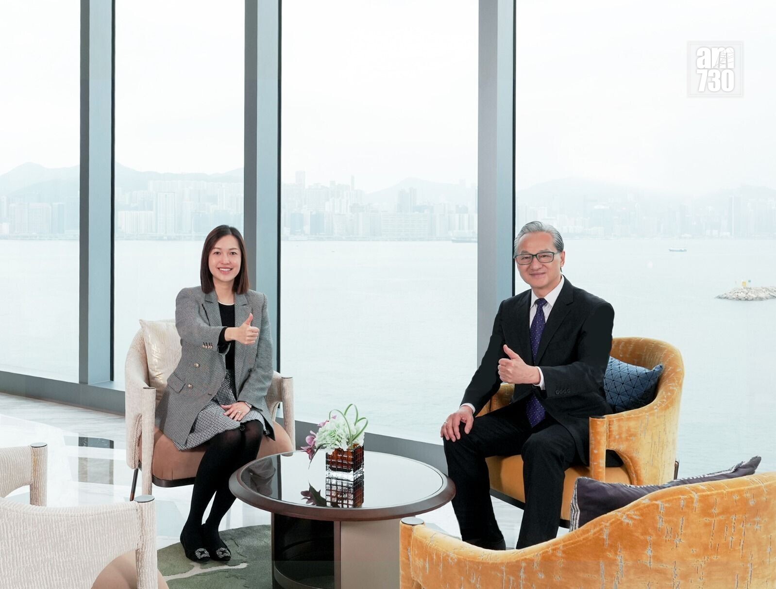 Two professionals, a woman and a man, sit in a bright lounge by large windows with a harbor view, giving thumbs up.