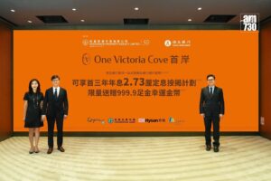 Three professionally dressed people stand in front of an orange promotional backdrop for One Victoria Cove with logos and Chinese text