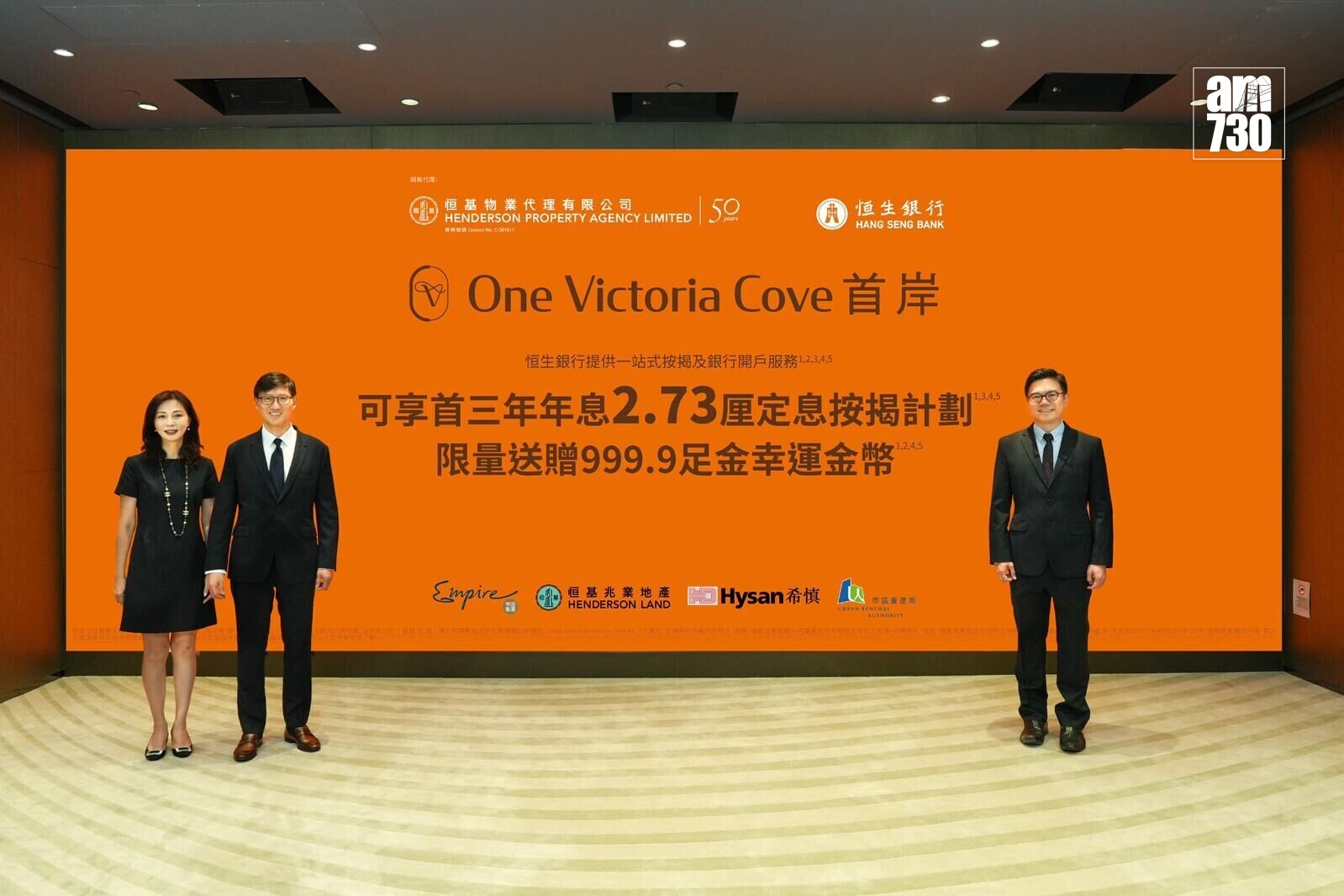 Three professionally dressed people stand in front of an orange promotional backdrop for One Victoria Cove with logos and Chinese text.