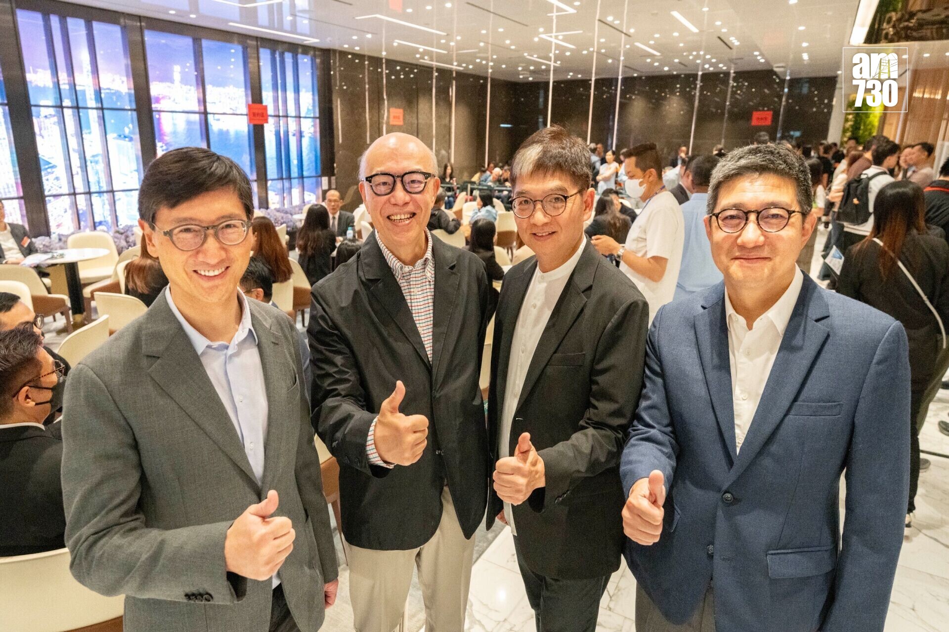 Four men in suits posing and giving thumbs up at a bustling indoor event with glass walls and tables in the background.