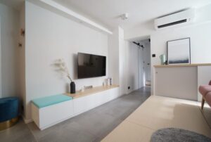 Bright modern living room with wall mounted TV white storage unit and pastel accents hallway to the right ceiling air conditioner above