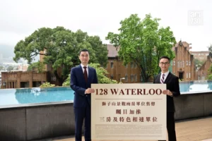 Two men in suits hold a large sign reading 128 WATERLOO outdoors by a pool with green trees and brick buildings in the background