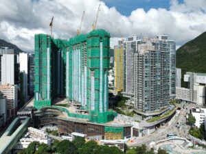 High rise construction site with green safety netting and multiple cranes against a cloudy sky
