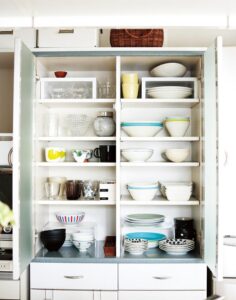 Open kitchen cabinet filled with assorted dishes bowls cups and glassware on white shelves