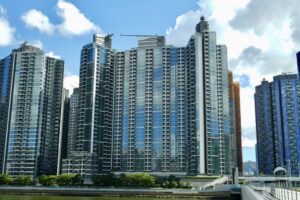 Row of tall glass residential towers along a waterfront under a blue sky with scattered clouds A promenade with trees runs along the water in the foreground