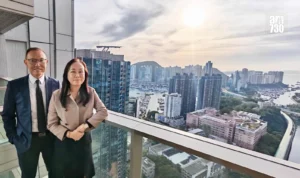 Two professionals pose on a glass balcony with a dense coastal city and marina behind them top right logo AM730 visible