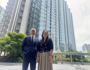Two professionally dressed people pose outside a glass fronted office building man in a navy suit woman in a blazer and beige skirt