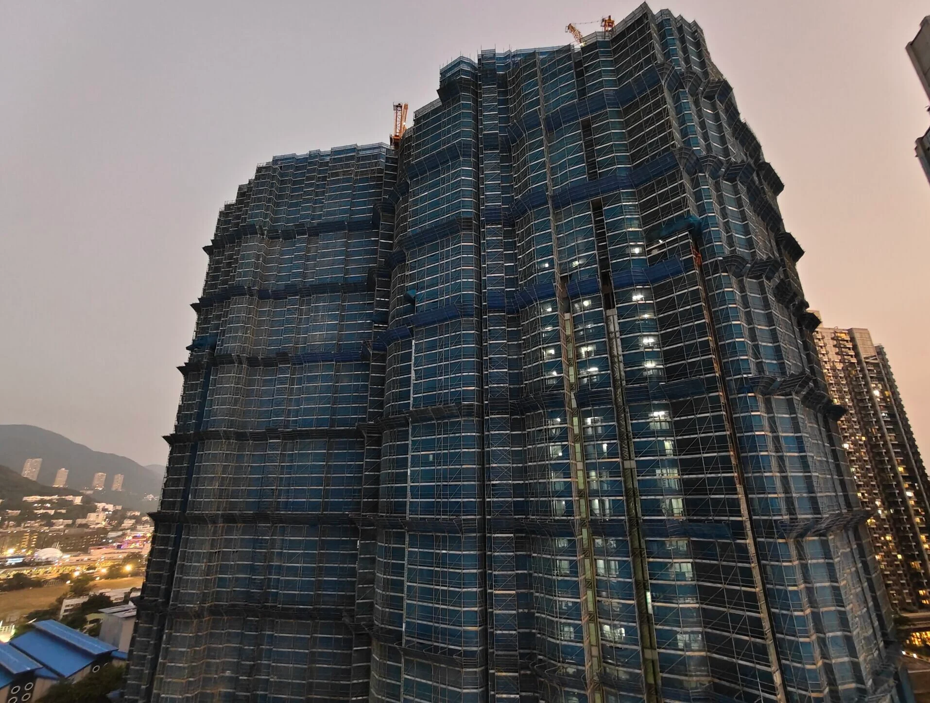 Under-construction blue-glass skyscraper with cranes on top and a city lit skyline at dusk.