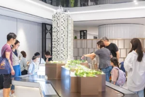 People gather around a large detailed scale model of a tall residential tower in a showroom or exhibition space examining the model closely