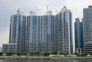 Row of blue glass high rise apartment buildings along a waterfront promenade with construction cranes on the rooftops and trees in front