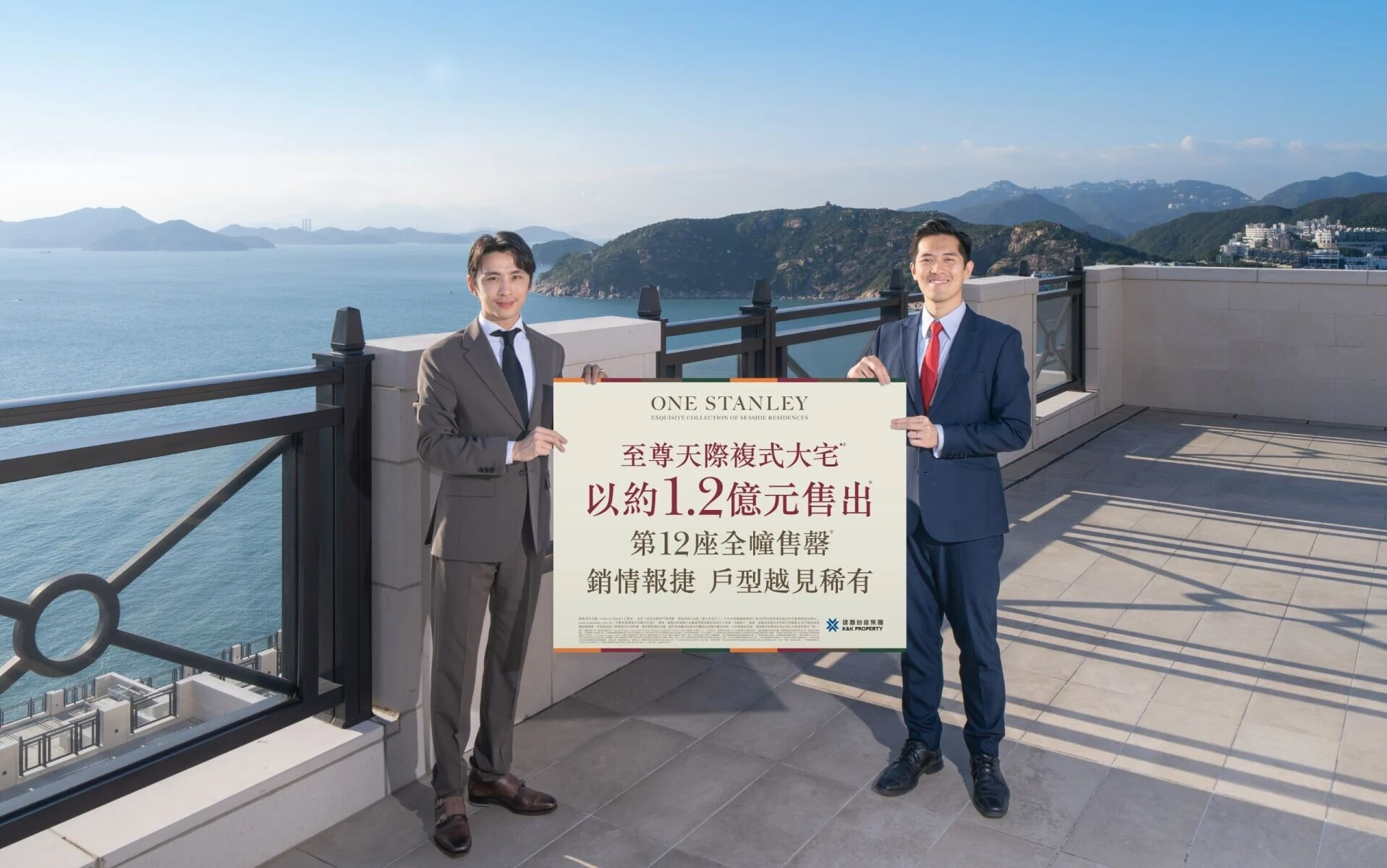 Two men in business suits stand on a seaside terrace, holding a large sign with Chinese text, ocean and mountains in the background.