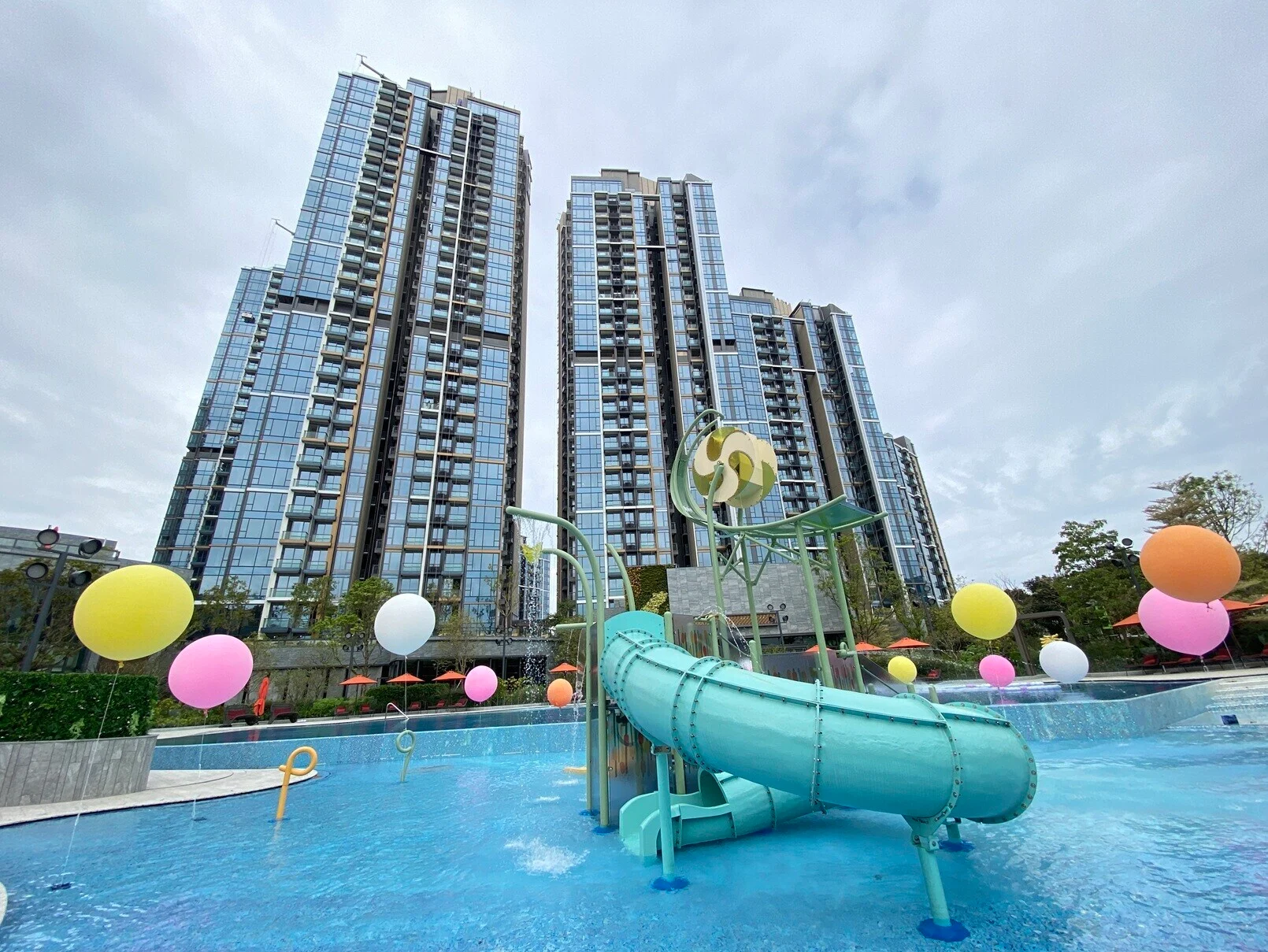 Tall glass residential towers with a turquoise waterslide in a pool area, surrounded by colorful balloons and amenities.