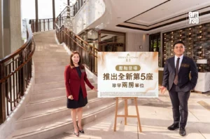 Two staff members in business attire pose beside a large signboard in a marble hotel lobby with a curved staircase behind them