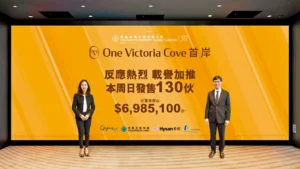 兩房698萬入場 首岸3期周日首輪賣130伙 | 利嘉閣地產有限公司 Two formally dressed presenters stand on either side of a large orange One Victoria Cove banner with Chinese text and the price 85100
