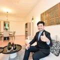 Man in a suit giving a thumbs up while seated on a beige sofa in a modern living room with a dining area in the background