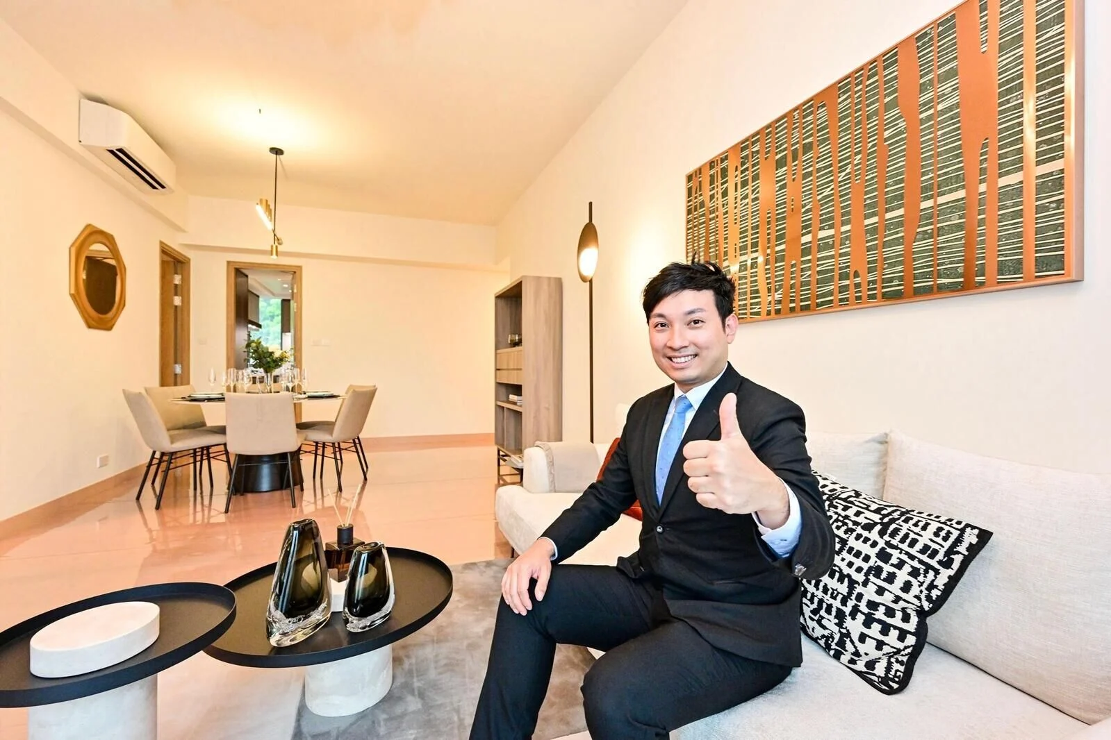 Man in a suit giving a thumbs up while seated on a beige sofa in a modern living room with a dining area in the background