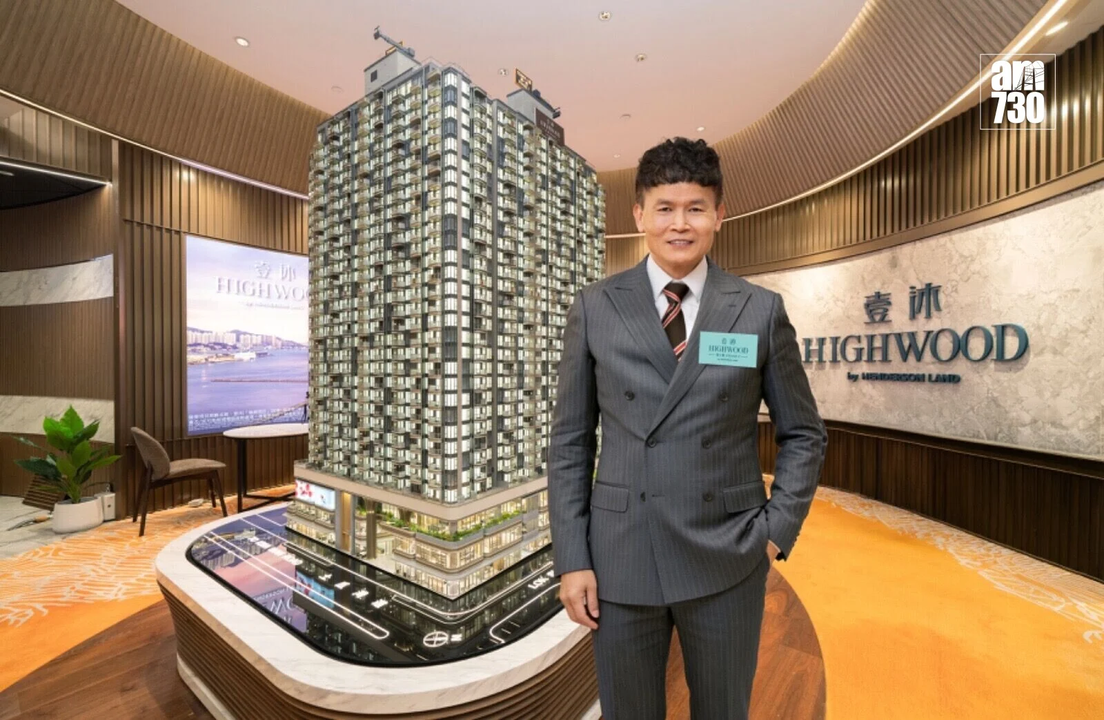 Man in a gray pinstripe suit stands beside a large scale model of a high-rise building in a Highwood development showroom. AM730 logo is visible in the corner.