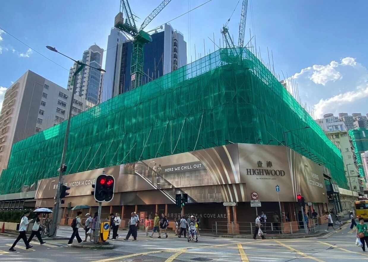 Construction site wrapped in green safety netting with cranes overhead beside a busy city street intersection, under construction.