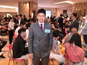 Man in a gray pinstripe suit with a name badge stands at the center of a crowded event hall filled with seated attendees and crowds in the background