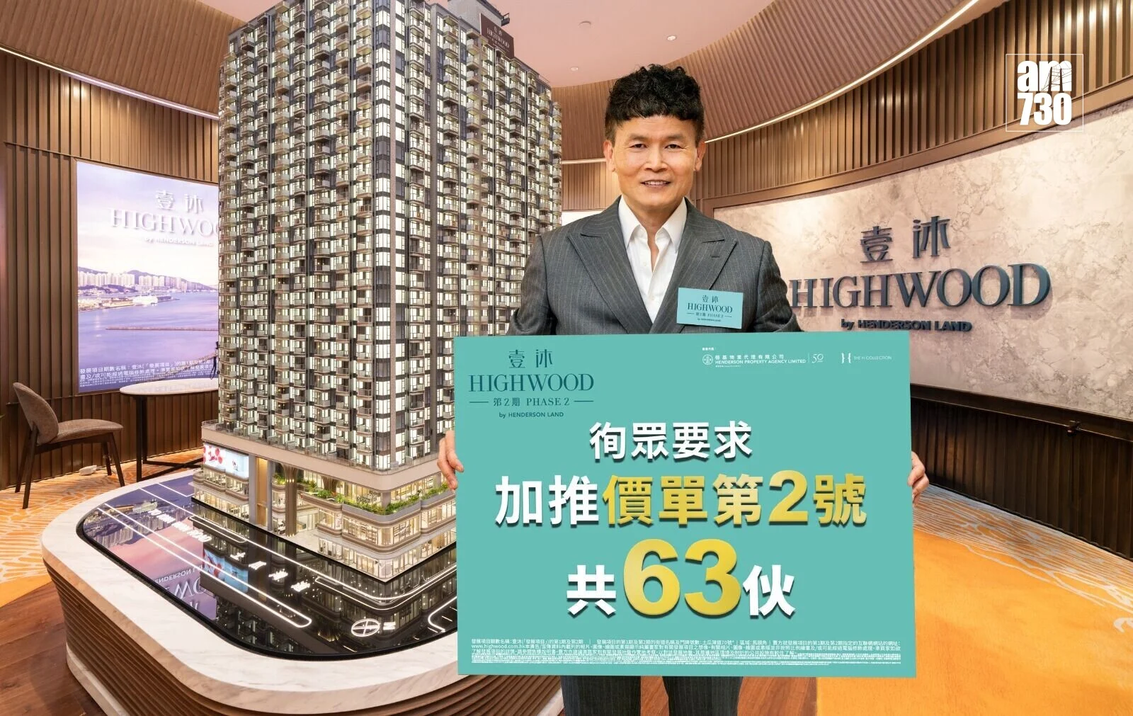 Smiling man in a suit holds a teal Highwood Phase 2 sign beside a detailed high-rise model in a showroom.
