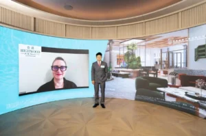 Man in a gray suit stands in a curved showroom backdrop a video call on the left shows a woman with glasses while a living room scene fills the right